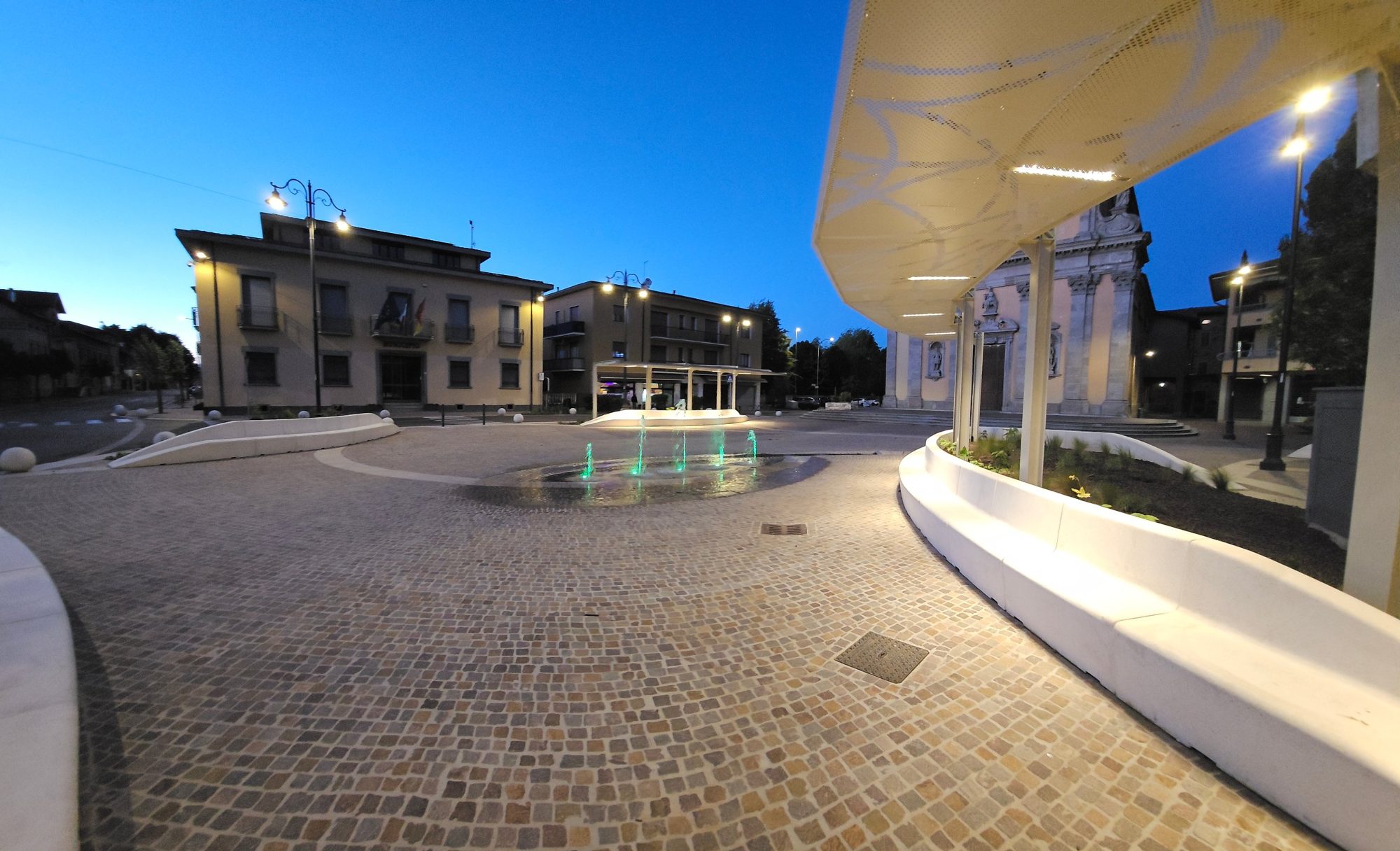 Piazza San Vittore la sera, con la fontana illuminata e i pergolati